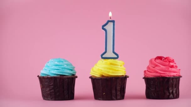 Cupcakes con crema y vela festiva encendida en forma de número uno sobre fondo rosa. Apague la vela en una magdalena festiva. Disparo en movimiento de primer plano — Vídeo de stock