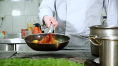 Mutfakta aşçı kızartma tavasında sebze kızartıyor, sebzeleri karıştırıyor. Aşçı tavada domates ve biber karıştırır. kamera çekim videosu çerçevesinde hareket eder