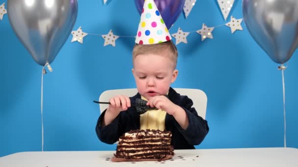 Menino feliz em uma jaqueta festiva com uma borboleta e um chapéu na cabeça come uma colher de bolo de chocolate em um fundo azul decorado com balões e estrelas. — Vídeo de Stock