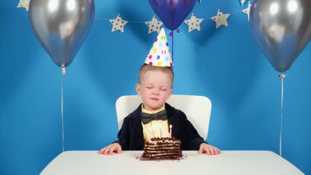 Feliz niño en la fiesta de cumpleaños cierra los ojos pide deseo y sopla velas en pastel de chocolate en el fondo festivo con globos y decoraciones hechas de estrellas. Cumpleaños del plan general — Vídeos de Stock