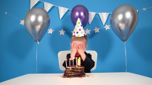 Menino feliz aniversário senta-se em uma mesa festiva em uma festa de aniversário, fecha os olhos, faz um desejo e apaga as velas, em seguida, bate palmas suas mãos. Em um fundo festivo azul com balões e fitas — Vídeo de Stock