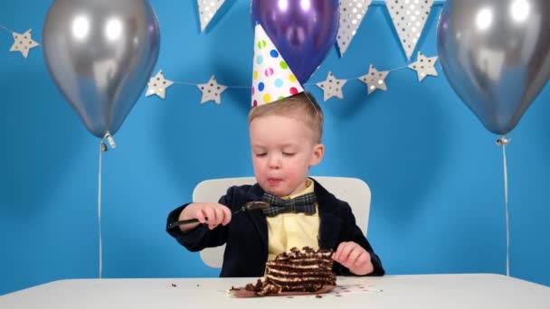 Niño pequeño está comiendo un pastel de chocolate dulce, su cara está untada con crema batida de pastel, los niños disfrutan comiendo pastel de cumpleaños. Sobre un fondo azul con decoraciones festivas de globos y estrellas — Vídeos de Stock