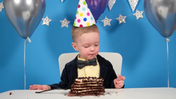 Feliz cumpleaños niño come una cucharada de pastel de chocolate, lame sus dedos manchados de postre, y se limpia la boca manchada con la manga de su chaqueta. concepto de fiesta y cumpleaños — Vídeos de Stock