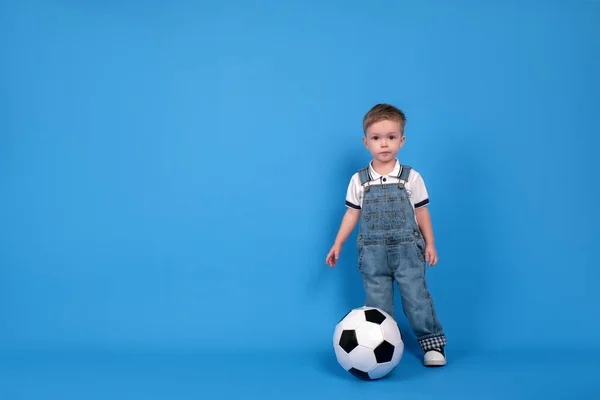 Um pequeno campeão de futebol. Adorável menino europeu com bola de futebol sentado no chão sobre fundo azul em estúdio, espaço de cópia, conceito de esporte — Fotografia de Stock