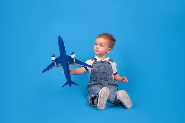 Happy child sits holding plane in his hand and imagining how he is flying on plane on blue background. Imagination, creativity and concept ideas. — Stock Photo, Image