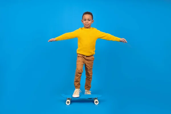 Boy African-American boy in everyday clothes skateboards having fun and smiling. Hands arranged for balancing on blue background with space for text. Concept of activity and happy childhood.