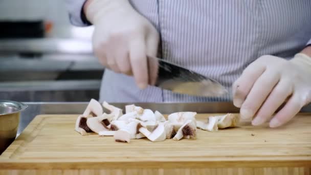 Ο σεφ του Καυκάσου στην κουζίνα του εστιατορίου με γάντια κόβει ωμά μανιτάρια με μαχαίρι σε ξύλο κοπής μπαμπού και τα βάζει σε μεταλλικό μπολ. Βίντεο διαφανειών Dolly — Αρχείο Βίντεο