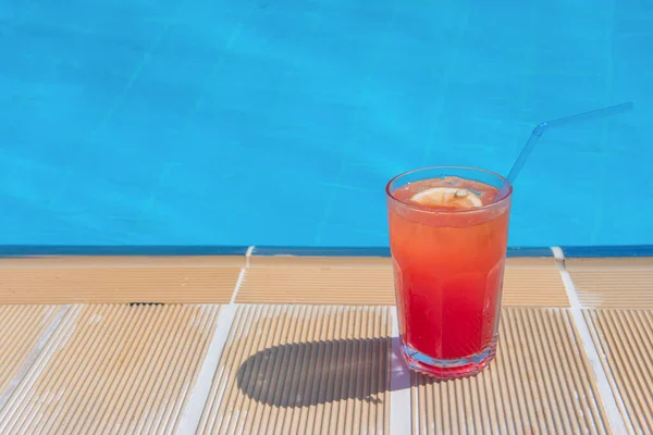 Fresh alcoholic red cocktail sex on beach with ice in glass on edge of pool in summer weather against background washed up pool and recreation area. Concept good holiday and travel, place for text