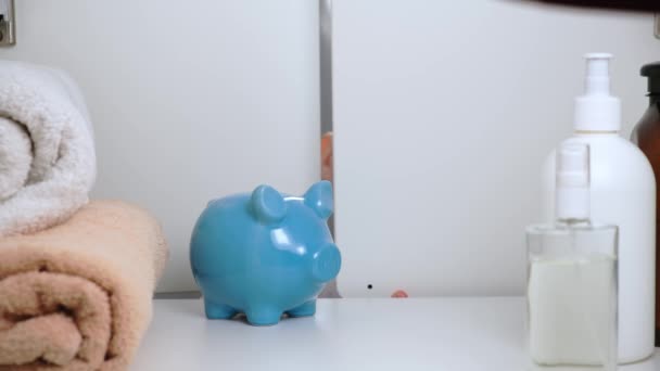 Menino loiro pequeno em t-shirt branca abre a porta do armário com toalhas e produtos de higiene no quarto e coloca dinheiro verde em piggy Bank azul. Kid coleta dinheiro para brinquedo ou presente. Vista de trás do Gabinete. — Vídeo de Stock