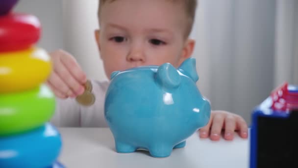 Der kleine blonde kaukasische Junge steckt Münzen und Geld in ein blaues Sparschwein aus Keramik mit Blick aus dem Kleiderschrank im Schrank des Kinderzimmers. Macht eine Investition sammelt Geld für die Zukunft, Spielzeug oder Geschenk. — Stockvideo