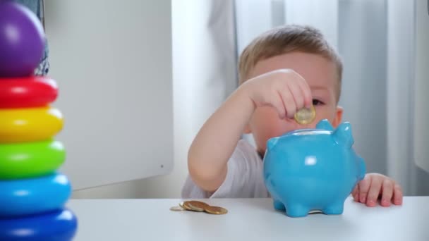 Der kleine blonde kaukasische Junge steckt Münzen und Geld in ein blaues Sparschwein aus Keramik mit Blick aus dem Kleiderschrank im Schrank des Kinderzimmers. Macht eine Investition sammelt Geld für die Zukunft, Spielzeug oder Geschenk. — Stockvideo