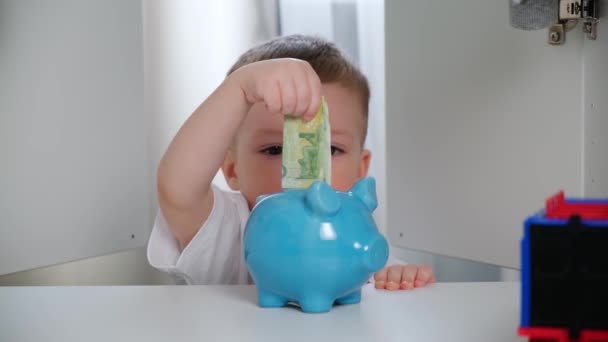 Malý roztomilý chlapec v bílém tričku se snaží dát zelené papírové peníze do modré keramické prasátko banky, peníze se nelepí a ohýbá. od první osoby. Pohled z vnitřní skříně. Investice peněz. — Stock video