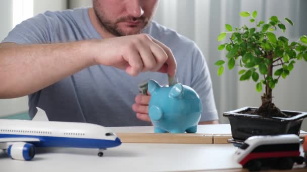 Un homme caucasien aux cheveux foncés dépose des euros et des pièces de monnaie dans une banque de piges bleues pour les épargner. Investit et économise pour l'avenir en vacances ou pour acheter une voiture ou un bien immobilier, sur fond de salon. — Video