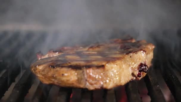 Köttbiffen ligger på en järngrill på varm kolgrill, kocken smetar olja på köttfilé och förvandlar stekt fläsk med tång, rök och flammor fyllning ram. Närbild video — Stockvideo