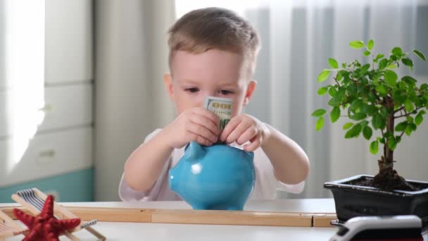Netter blonder kaukasischer Junge spielt im Kinderzimmer, steckt Papiergeld in blaues Keramik-Sparschwein, sammelt Geld für Erholung vor dem Hintergrund eines Tisches mit Eisenbahn, Seesternen und grünem Bonsaibaum. — Stockvideo