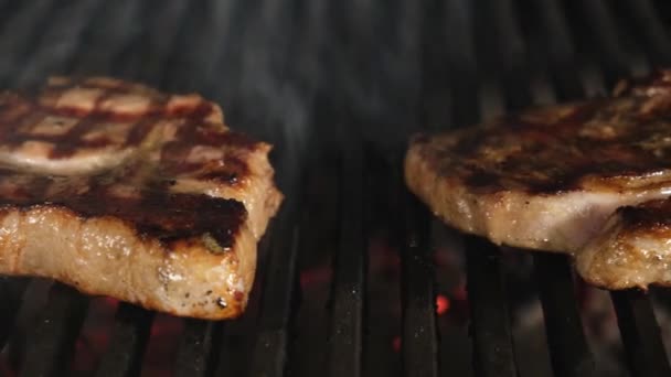 Deux steaks de viande juteux à la croûte dorée et des lamelles de grill barbecue sont préparés sur une flamme ouverte de grill barbecue, cuire la viande tournante avec des pinces métalliques filet de porc. Vidéo coulissante Dolly. — Video
