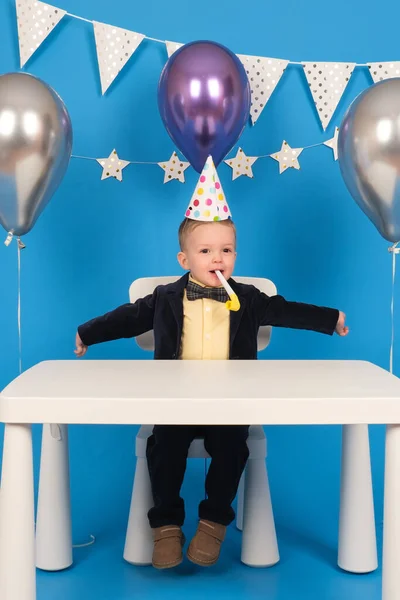 Boldog szőke fiú ül az asztalnál ünnepi kalapban születésnapi party, fújja ünnepi síp öröm és felemeli a kezét oldalra, ünnepli elérése cél, a díszített kék háttér. — Stock Fotó