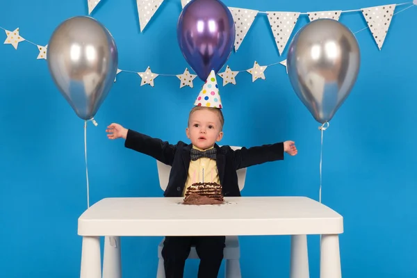 Boldog kaukázusi fiú ül asztalnál ünnepi kúp alakú kalap születésnapi party, örül, és felemeli a kezét oldalra, ünnepli győzelem és a cél elérése, a díszített kék háttér. — Stock Fotó