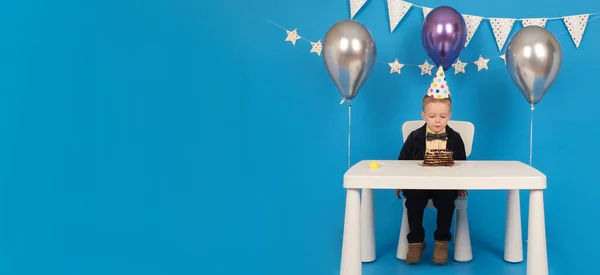 Happy blond boy sits at table in festive hat cone on birthday blows out colored candles on chocolate cake, make wish, receive gifts. on decorated blue background with balloons, stars and flags. — Stock Photo, Image