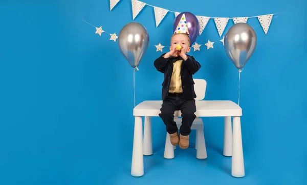 Feliz chico caucasiano rubio está sentado sobre la mesa en sombrero festivo, soplando alegremente silbato festivo y extendiendo sus brazos a los lados, celebrando el logro de la meta, sobre fondo azul decorado.. — Foto de Stock