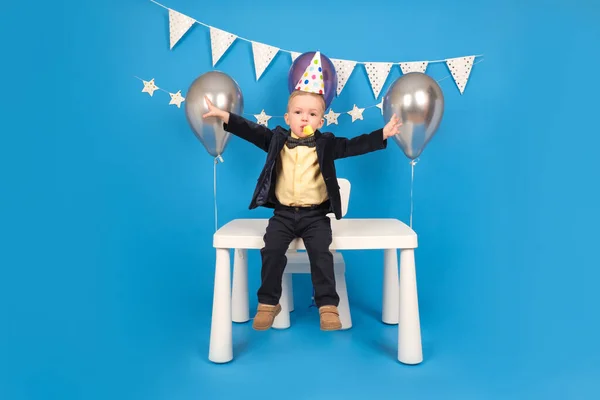Boldog szőke fiú ül az asztalon ünnepi kúp kalap a születésnapján, örül, és felemeli a kezét oldalra ünnepli győzelem és a cél elérése, kívánság, a díszített kék háttér. — Stock Fotó