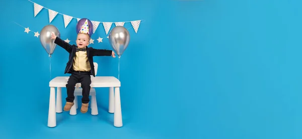 Feliz negrito está sentado sobre la mesa en un sombrero de cono festivo en su cumpleaños, regocijándose y levantándose las manos para celebrar la victoria y alcanzar la meta, haciendo deseos, sobre fondo azul decorado.. — Foto de Stock