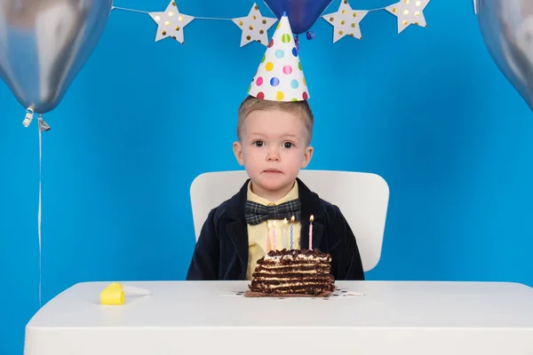 Feliz menino loiro senta-se à mesa em cone chapéu festivo no aniversário sopra velas coloridas no bolo de chocolate, fazer desejo, receber presentes. em fundo azul decorado com balões, estrelas e bandeiras. — Fotografia de Stock