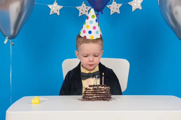 El niño rubio feliz se sienta a la mesa en el cono festivo del sombrero durante el cumpleaños sopla velas coloreadas en el pastel de chocolate, haga el deseo, reciba regalos. sobre fondo azul decorado con globos, estrellas y banderas.. — Foto de Stock