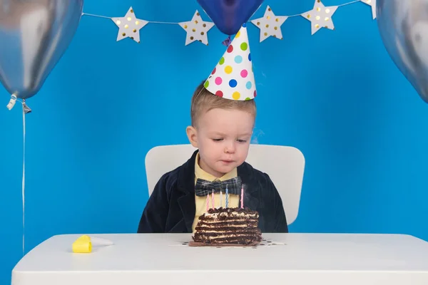 El niño rubio feliz se sienta a la mesa en el cono festivo del sombrero durante el cumpleaños sopla velas coloreadas en el pastel de chocolate, haga el deseo, reciba regalos. sobre fondo azul decorado con globos, estrellas y banderas.. — Foto de Stock