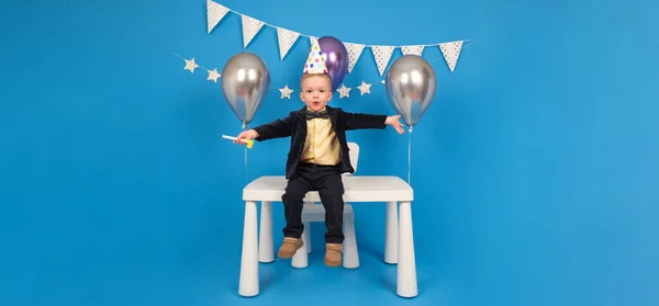 Feliz negrito está sentado sobre la mesa en un sombrero de cono festivo en su cumpleaños, regocijándose y levantándose las manos para celebrar la victoria y alcanzar la meta, haciendo deseos, sobre fondo azul decorado.. — Foto de Stock