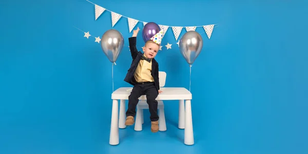 Feliz negrito está sentado sobre la mesa en un sombrero de cono festivo en su cumpleaños, regocijándose y levantándose las manos para celebrar la victoria y alcanzar la meta, haciendo deseos, sobre fondo azul decorado.. — Foto de Stock