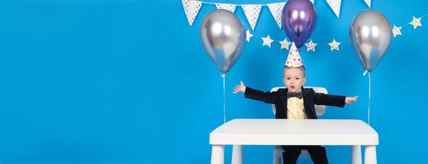 Feliz chico caucásico se sienta a la mesa en un sombrero festivo en forma de cono en la fiesta de cumpleaños, regocijándose y levantando las manos a un lado, celebrando la victoria y alcanzando la meta, sobre fondo azul decorado.. — Foto de Stock