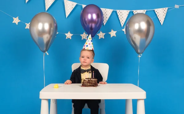 Happy blond Caucasian boy in festive hat, sitting at table with chocolate cake celebrates his birthday on blue background decorated with balloons, stars and flags. — Stock Photo, Image