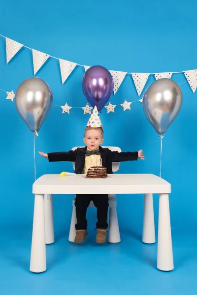 Boldog kaukázusi fiú ül asztalnál ünnepi kúp alakú kalap születésnapi party, örül, és felemeli a kezét oldalra, ünnepli győzelem és a cél elérése, a díszített kék háttér. — Stock Fotó