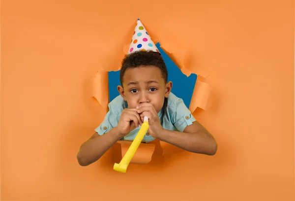 Happy african american boy blows birthday pipe with cone-shaped cap on his head against peach-colored ragged background of torn paper edges. Childrens fun concept. Kids celebrate their birthday. — Stock Photo, Image