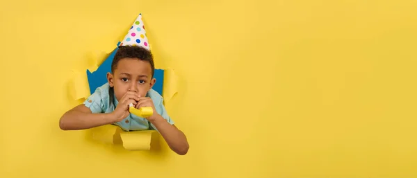 Menino afro-americano feliz soprando tubo festivo na festa de aniversário com um boné de cone na cabeça em um fundo rasgado amarelo bordas rasgadas de papel. Crianças conceito divertido. O miúdo está a celebrar a festa de aniversário.. — Fotografia de Stock
