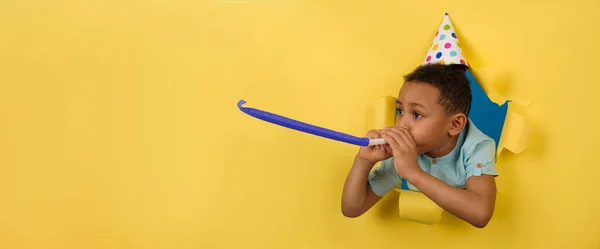 Feliz chico afroamericano soplando pipa festiva en la fiesta de cumpleaños con una tapa de cono en la cabeza sobre un fondo amarillo roto bordes rotos de papel. Concepto divertido para niños. El chico está celebrando la fiesta de cumpleaños.. — Foto de Stock