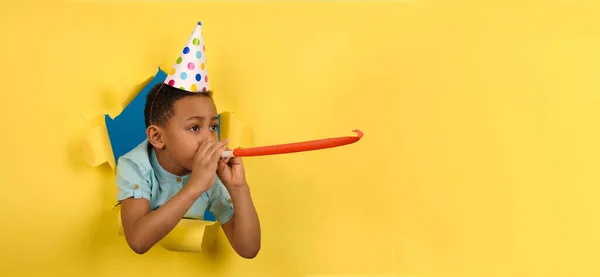 Feliz chico afroamericano soplando pipa festiva en la fiesta de cumpleaños con una tapa de cono en la cabeza sobre un fondo amarillo roto bordes rotos de papel. Concepto divertido para niños. El chico está celebrando la fiesta de cumpleaños.. —  Fotos de Stock