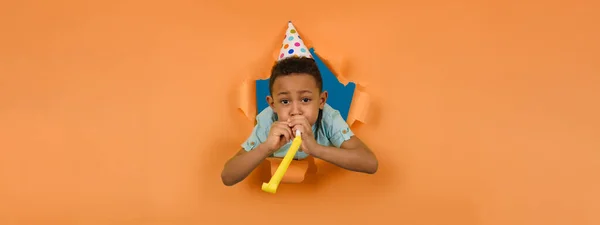 Happy african American boy blows birthday pipe with cone-shaped cap on his head against peach-colored ragged background of torn paper edges. El concepto de diversión infantil. Los niños celebran su cumpleaños. — Foto de Stock