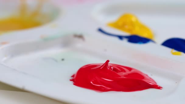 Close-up artista aprieta la pintura fuera del tubo y mezcla dos pinturas acrílicas en la paleta de color rojo y verde para obtener el color marrón. Dibuja una imagen en un estudio de arte sobre lienzo . — Vídeos de Stock