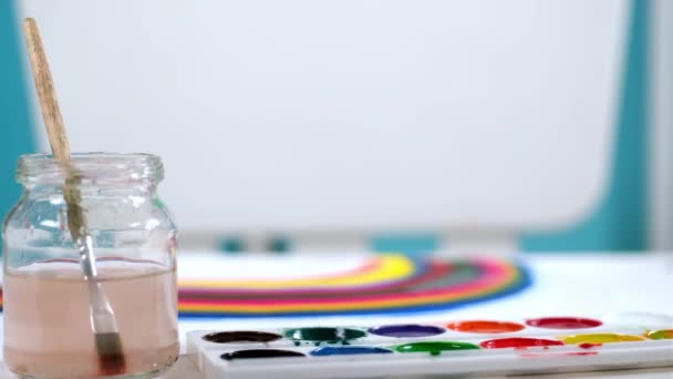 Maler nimmt Nahaufnahme Pinsel und trägt blaue Aquarellfarbe auf Palette, Zeichnung Regenbogen auf verschwommenem Hintergrund. Künstler zeichnet Bild auf Leinwand in Art Studio. Regenbogen ist Symbol des Sieges über das Virus. — Stockvideo