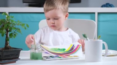 Çocuk odasındaki sarışın beyaz erkek sanatçı, suluboya ile gökkuşağı çizmiş ve ailesine resim çizmiş, tuvalin arkasından yukarı doğru bakmış ve eğlenmiş. Gökkuşağı virüse karşı zaferin sembolüdür.