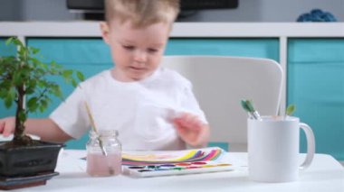 Çocuk odasındaki beyaz çocuk ressam tablo malzemeleriyle masaya oturur, fırça alır ve gökkuşağının suluboya resmini çizer. Gökkuşağı, Coronavirus 'a karşı zafer ve umudun sembolüdür.