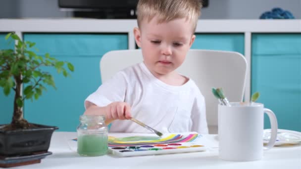 Blonde Καυκάσιος καλλιτέχνης αγόρι στο παιδικό δωμάτιο αντλεί πράσινο γρασίδι στο τραπέζι με ακουαρέλες, το παιδί βρέχει το πινέλο στο νερό, εφαρμόζοντας χρώμα για να βουρτσίσετε και σχέδιο σε χαρτί — Αρχείο Βίντεο