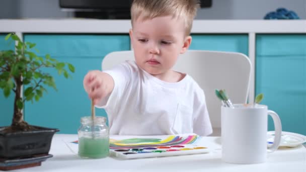 Loira caucasiano menino artista na sala das crianças desenha grama verde à mesa com aquarelas, criança molhando o pincel na água, aplicando tinta para escovar e desenho em papel — Vídeo de Stock