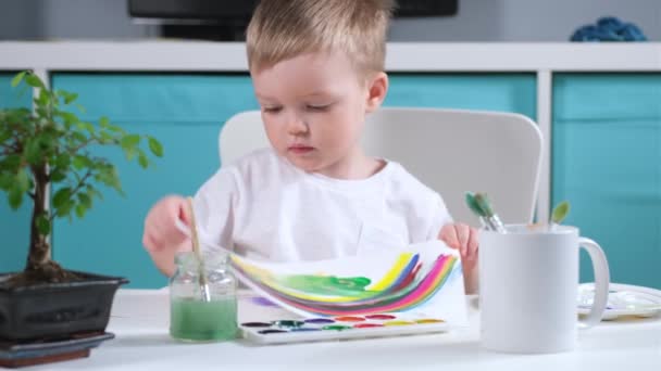 Blonder kaukasischer Junge Künstler im Kinderzimmer zeichnete Regenbogen mit Aquarellen und zeigt Zeichnung zu seinen Eltern, hebt oben schaut aus hinter Leinwand und hat Spaß. Regenbogen ist Symbol des Sieges über das Virus — Stockvideo