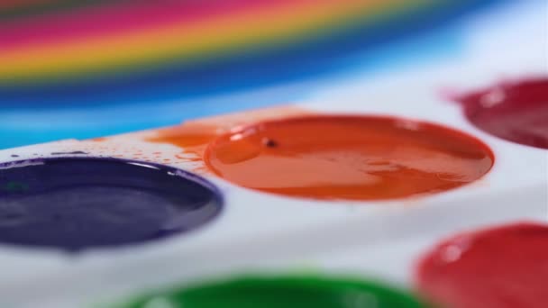 Närbild målare tar borste och tillämpar röd akvarell färg på paletten, drar regnbåge på suddig bakgrund. En konstnär ritar bild på duk i en konststudio. — Stockvideo