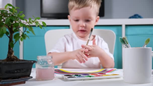 Rubio caucásico niño artista dibuja arco iris en habitación de los niños en la mesa con acuarelas, cepillo mojado niño en agua, aplicando pintura sobre el pincel y dibujo sobre papel. arco iris símbolo de esperanza contra el virus — Vídeo de stock