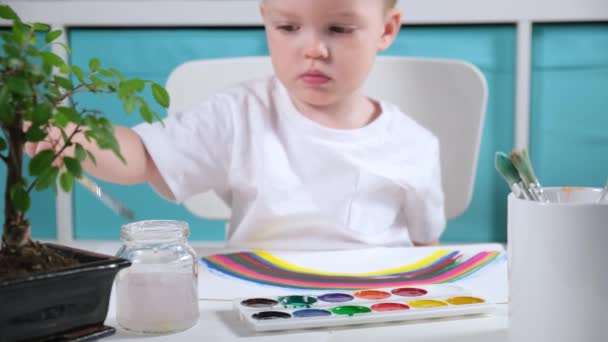 Loira caucasiano menino artista desenha arco-íris na sala das crianças à mesa com aquarelas, criança molhando escova na água, aplicando tinta no pincel e desenho em papel. arco-íris símbolo de esperança contra o vírus — Vídeo de Stock