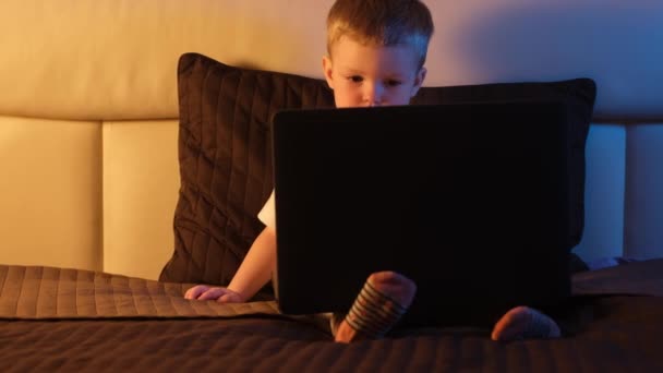 Leuke blonde blanke jongen alleen in de slaapkamer 's nachts zitten op bed, typen op laptop toetsenbord, surfen of trawlen internet. Nachtlampje in de kamer, maanlicht door het raam. Dolly glijdt door video. — Stockvideo